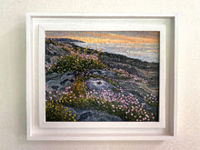 Load image into Gallery viewer, A framed coastal landscape painting hanging on a light beige wall. The artwork depicts rocky cliffs covered with pink and yellow wildflowers, overlooking a calm ocean at sunset.