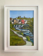 Load image into Gallery viewer, A framed landscape painting of a scenic village with a waterfall, green hills, and a blue sky, presented in a white wooden frame against a light beige wall.