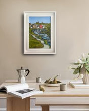 Load image into Gallery viewer, A framed landscape art print depicting a scenic coastal village with green hills, blue water, and red-roofed buildings, displayed on a beige wall above a wooden table with coffee cups and books.