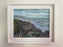 Load image into Gallery viewer, A framed coastal landscape painting hanging on a light beige wall. The artwork depicts rocky shoreline, ocean water, distant land, and a cloudy sky, enclosed in a white frame with a white mat.